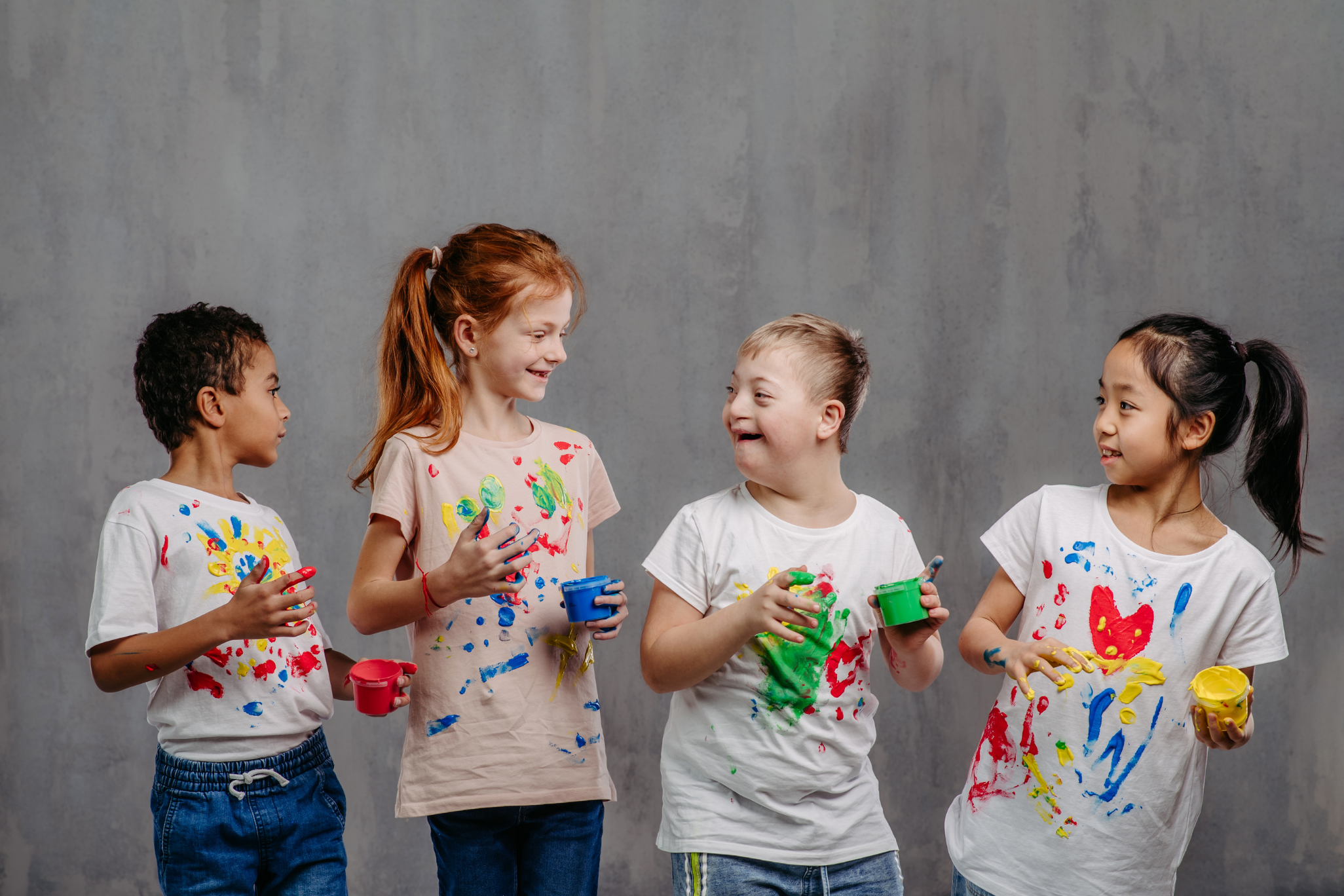 Four children joyfully hold paint pots, their shirts splattered with vibrant colors, against a gray background, conveying creativity and friendship.
