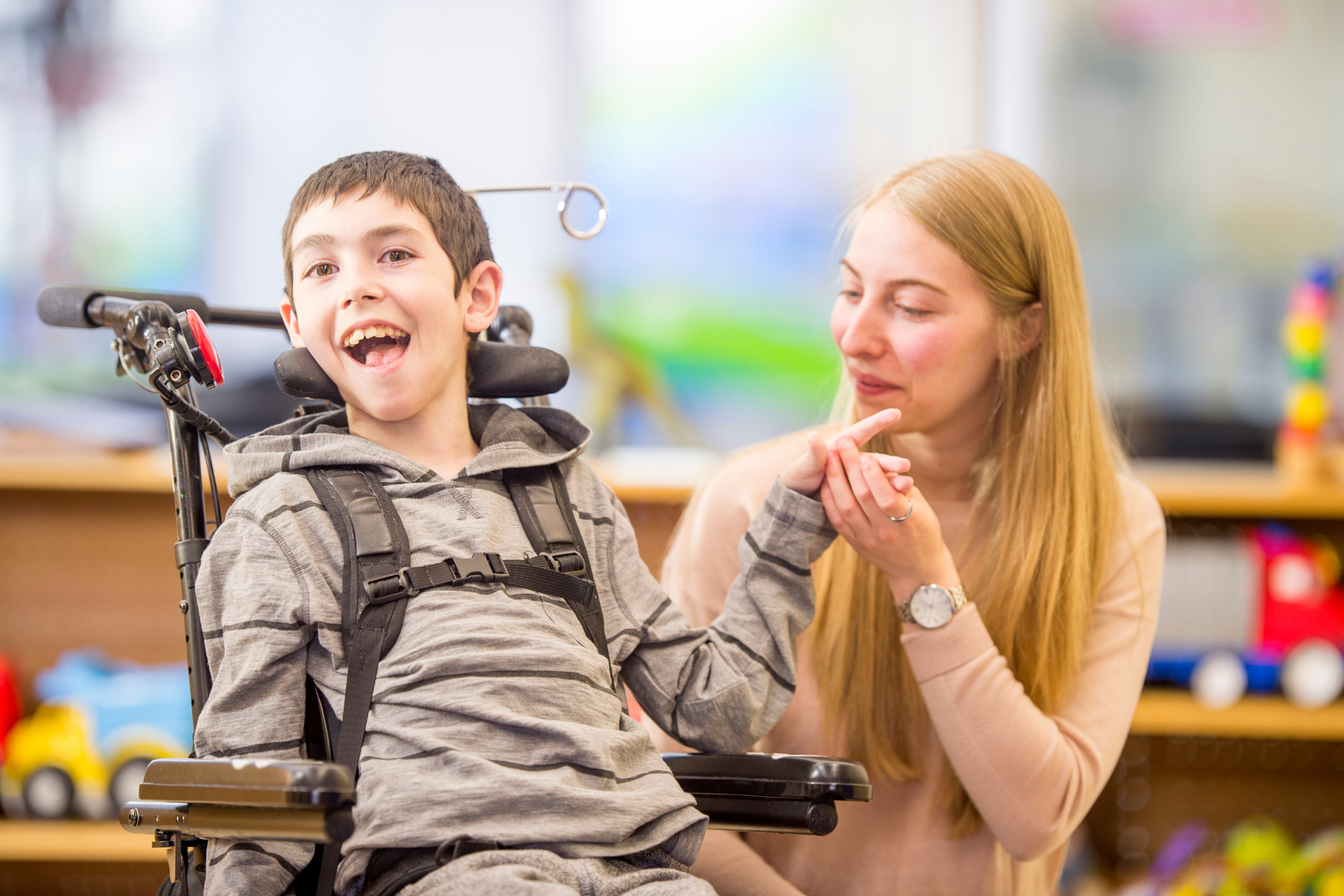 A smiling boy in a gray hoodie sits in a wheelchair, joyfully interacting with a woman beside him in a classroom setting. The mood is warm and supportive.